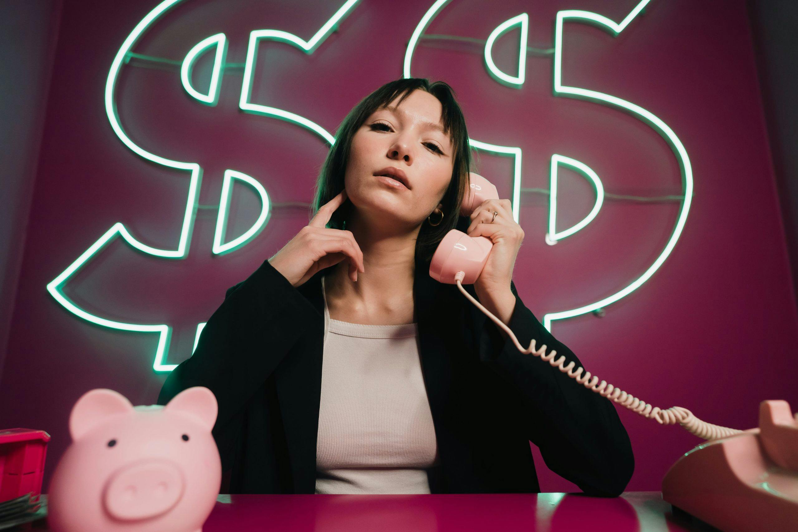 A woman taking a call on a pink telephone. There is a piggy bank on the desk and dollar signs in the background.