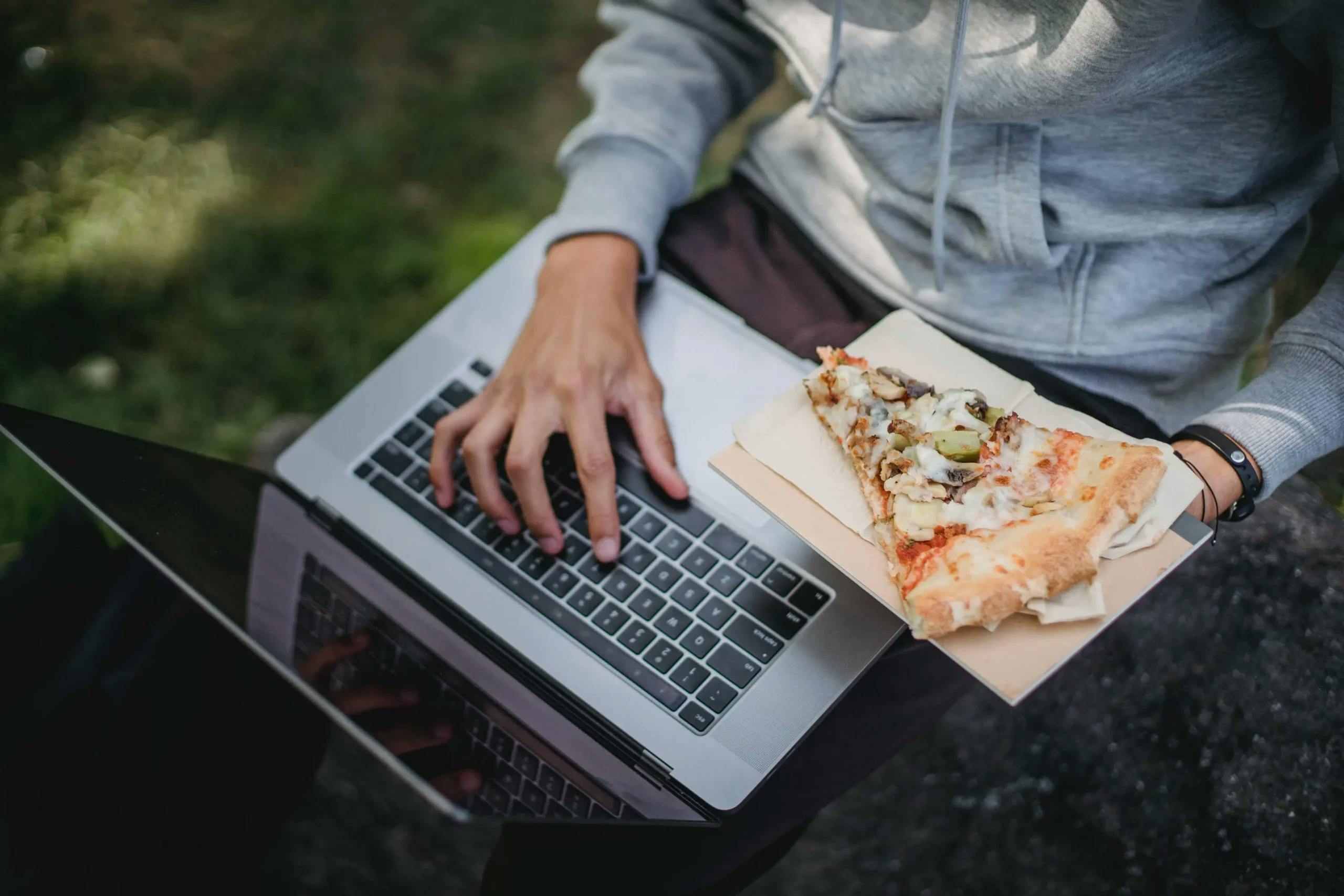 A young person working on a laptop while eating a slice of pizza.